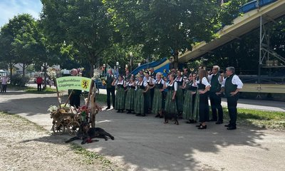 Eine Musikgruppe in traditioneller Kleidung steht in einem Park neben einem Schild mit der Aufschrift "Jagdhornbläser Göppingen seit 1965". Einige Hunde liegen entspannt am Boden.