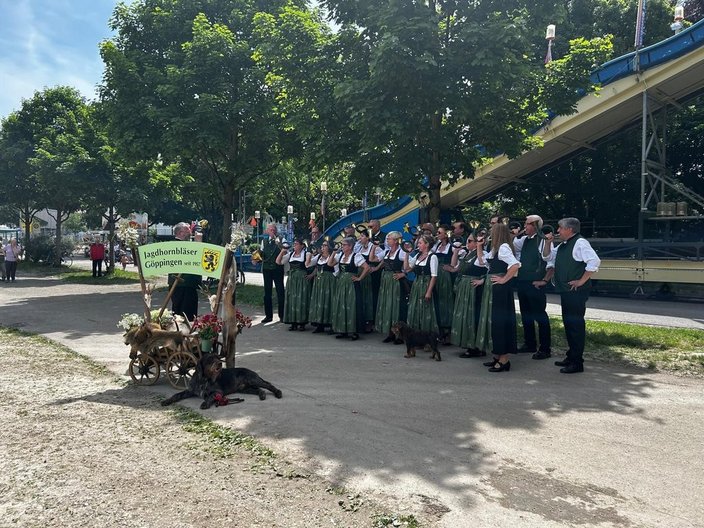 Eine Musikgruppe in traditioneller Kleidung steht in einem Park neben einem Schild mit der Aufschrift "Jagdhornbläser Göppingen seit 1965". Einige Hunde liegen entspannt am Boden.