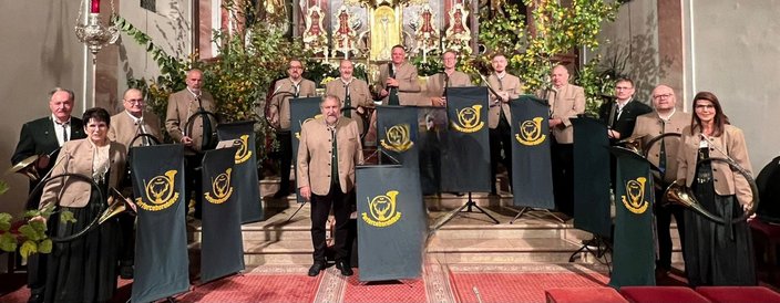 Menschen in traditionellen Trachten stehen mit Alphörnern vor einem Altar. Im Hintergrund geschmückter Altar mit Pflanzen. Text auf Stoffen: "Parforcehornbläser". Die Szene findet in einer Kirche statt.