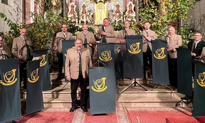Menschen in traditionellen Trachten stehen mit Alphörnern vor einem Altar. Im Hintergrund geschmückter Altar mit Pflanzen. Text auf Stoffen: "Parforcehornbläser". Die Szene findet in einer Kirche statt.